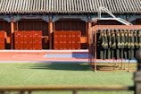 A courtyard with a traditional tiled roof structure, featuring elaborate decorative elements on the beams. In the courtyard, there are several brown lockers neatly arranged near the wall. In the foreground, a rack holds numerous black uniforms with yellow stripes and red insignias. The ground is divided into a green artificial grass area and a red and blue outdoor court, likely for sports.
