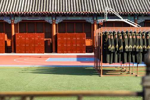 A courtyard with a traditional tiled roof structure, featuring elaborate decorative elements on the beams. In the courtyard, there are several brown lockers neatly arranged near the wall. In the foreground, a rack holds numerous black uniforms with yellow stripes and red insignias. The ground is divided into a green artificial grass area and a red and blue outdoor court, likely for sports.
