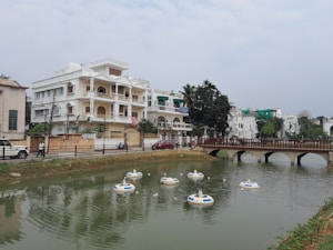 A residential area featuring large white buildings with multiple stories and balconies is situated beside a small body of water. Several floating devices are visible in the water, and a wooden bridge spans across it. The scene includes trees and some greenery surrounding the buildings, along with parked vehicles and a few pedestrians on the street.