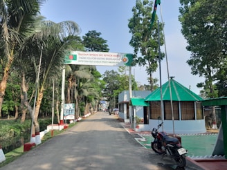 A well-maintained road leading to an integrated border check post. Trees are lined up along the sides, and buildings with green roofs are visible to the right. There is a large sign overhead that welcomes and thanks visitors to Bangladesh. A motorcycle is parked near the right side of the image.