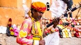 A man dressed in colorful traditional attire is playing a stringed musical instrument. In the background, there is a vibrant display of assorted sandals and shoes lined up against a wall in an outdoor setting.