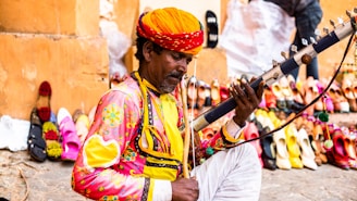A man dressed in colorful traditional attire is playing a stringed musical instrument. In the background, there is a vibrant display of assorted sandals and shoes lined up against a wall in an outdoor setting.