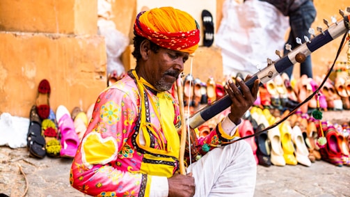 A man dressed in colorful traditional attire is playing a stringed musical instrument. In the background, there is a vibrant display of assorted sandals and shoes lined up against a wall in an outdoor setting.