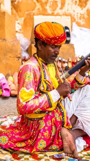 A person dressed in traditional, colorful attire is sitting on a patterned mat, playing a musical instrument. They wear a vibrant orange turban and a bright pink shirt adorned with intricate floral designs. Around them, various footwear items like shoes and sandals are displayed against a warm-colored wall.