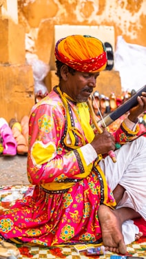 A person dressed in traditional, colorful attire is sitting on a patterned mat, playing a musical instrument. They wear a vibrant orange turban and a bright pink shirt adorned with intricate floral designs. Around them, various footwear items like shoes and sandals are displayed against a warm-colored wall.