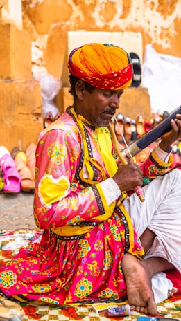 A person dressed in traditional, colorful attire is sitting on a patterned mat, playing a musical instrument. They wear a vibrant orange turban and a bright pink shirt adorned with intricate floral designs. Around them, various footwear items like shoes and sandals are displayed against a warm-colored wall.