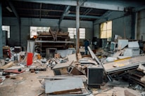 A cluttered and abandoned industrial space with piles of debris, broken furniture, and various discarded materials scattered across the floor. The walls are lined with large windows allowing some light to enter, highlighting the neglected state of the interior. A lone person is visible in the background, walking amidst the chaos.