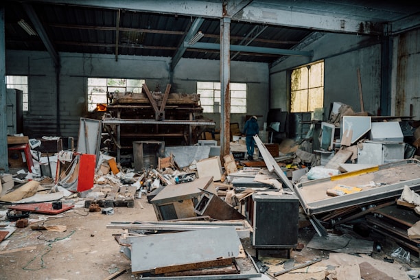 A cluttered and abandoned industrial space with piles of debris, broken furniture, and various discarded materials scattered across the floor. The walls are lined with large windows allowing some light to enter, highlighting the neglected state of the interior. A lone person is visible in the background, walking amidst the chaos.