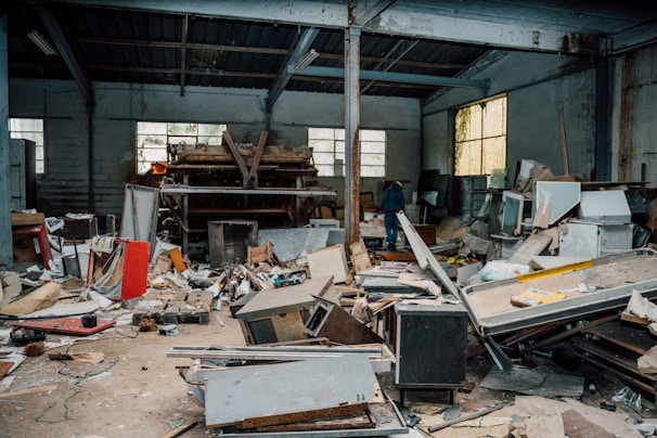 A cluttered and abandoned industrial space with piles of debris, broken furniture, and various discarded materials scattered across the floor. The walls are lined with large windows allowing some light to enter, highlighting the neglected state of the interior. A lone person is visible in the background, walking amidst the chaos.