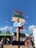 Colorful signpost with directional arrows pointing to different attractions such as Haven, Pier, Sea Life, Strand, and Reuzenrad. The background features a vibrant street scene with people walking and interacting near food stalls. The sky is clear with a few clouds, indicating a sunny day.