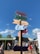 Colorful signpost with directional arrows pointing to different attractions such as Haven, Pier, Sea Life, Strand, and Reuzenrad. The background features a vibrant street scene with people walking and interacting near food stalls. The sky is clear with a few clouds, indicating a sunny day.