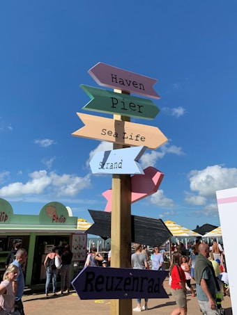 Colorful signpost with directional arrows pointing to different attractions such as Haven, Pier, Sea Life, Strand, and Reuzenrad. The background features a vibrant street scene with people walking and interacting near food stalls. The sky is clear with a few clouds, indicating a sunny day.