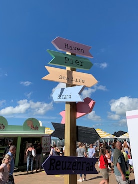 Colorful signpost with directional arrows pointing to different attractions such as Haven, Pier, Sea Life, Strand, and Reuzenrad. The background features a vibrant street scene with people walking and interacting near food stalls. The sky is clear with a few clouds, indicating a sunny day.