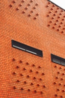 A brick wall with a pattern of protruding brick elements arranged in geometric shapes. Two rectangular windows with dark frames are inset into the wall, adding contrast to the orange-red of the bricks.