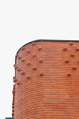 A red brick wall with a curved corner and protruding bricks against a plain white sky. The bricks are evenly laid with some bricks intentionally jutting out for decorative purposes. The structure appears to be well-maintained and clean.