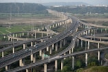 A complex network of elevated highways with multiple lanes and interchanges, surrounded by green fields and hills in the background. Cars and trucks are visible traveling on the roads, and the sky appears clear with a hint of an urban area on the horizon.