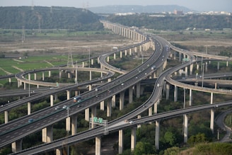 A complex network of elevated highways with multiple lanes and interchanges, surrounded by green fields and hills in the background. Cars and trucks are visible traveling on the roads, and the sky appears clear with a hint of an urban area on the horizon.