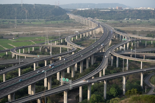 A complex network of elevated highways with multiple lanes and interchanges, surrounded by green fields and hills in the background. Cars and trucks are visible traveling on the roads, and the sky appears clear with a hint of an urban area on the horizon.