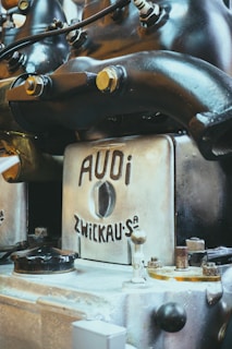 A close-up view of an antique or vintage engine with a metallic finish, featuring branding that reads 'AUDI ZWICKAU.' The machine components, including bolts and pipes, suggest an industrial or mechanical setting.