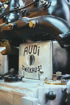 A close-up view of an antique or vintage engine with a metallic finish, featuring branding that reads 'AUDI ZWICKAU.' The machine components, including bolts and pipes, suggest an industrial or mechanical setting.