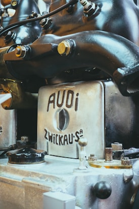 A close-up view of an antique or vintage engine with a metallic finish, featuring branding that reads 'AUDI ZWICKAU.' The machine components, including bolts and pipes, suggest an industrial or mechanical setting.