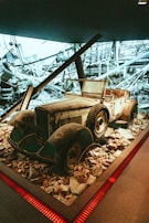 A vintage, rusted car is surrounded by rubble and debris indoors, under dim lighting. The background contains a dramatic black-and-white photograph of a destroyed industrial setting, enhancing the atmosphere of neglect and destruction.