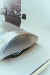 A sleek, silver, aerodynamic car design with a smooth, rounded rear section is on display. The setting appears to be a museum or exhibition, with informational panels and black-and-white photographs visible in the background, providing a historical context.