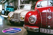 A showroom featuring vintage cars, prominently displaying a red IFA car alongside a beige car with round headlights and classic design elements. A large blue vintage truck is visible in the background. The setting is well-lit, with polished floors and several cars lined up for display.