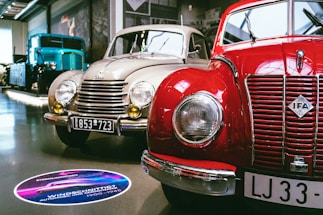 A showroom featuring vintage cars, prominently displaying a red IFA car alongside a beige car with round headlights and classic design elements. A large blue vintage truck is visible in the background. The setting is well-lit, with polished floors and several cars lined up for display.