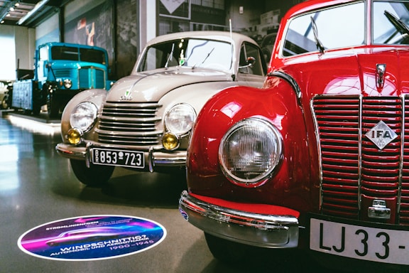 A showroom featuring vintage cars, prominently displaying a red IFA car alongside a beige car with round headlights and classic design elements. A large blue vintage truck is visible in the background. The setting is well-lit, with polished floors and several cars lined up for display.