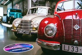 A showroom featuring vintage cars, prominently displaying a red IFA car alongside a beige car with round headlights and classic design elements. A large blue vintage truck is visible in the background. The setting is well-lit, with polished floors and several cars lined up for display.