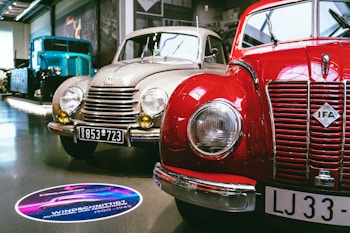 A showroom featuring vintage cars, prominently displaying a red IFA car alongside a beige car with round headlights and classic design elements. A large blue vintage truck is visible in the background. The setting is well-lit, with polished floors and several cars lined up for display.