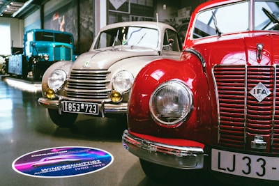 A showroom featuring vintage cars, prominently displaying a red IFA car alongside a beige car with round headlights and classic design elements. A large blue vintage truck is visible in the background. The setting is well-lit, with polished floors and several cars lined up for display.