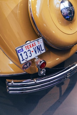 A vintage yellow car displays a Texas license plate with the number 133 VNL. The rear features chrome details, including a bumper and a round emblem. A classic design is highlighted by a polished metal surface, red lighting, and decorative elements.