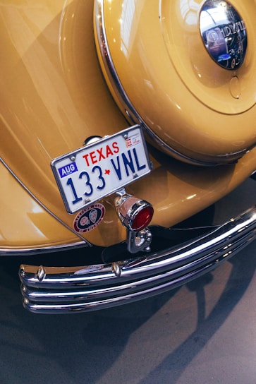 A vintage yellow car displays a Texas license plate with the number 133 VNL. The rear features chrome details, including a bumper and a round emblem. A classic design is highlighted by a polished metal surface, red lighting, and decorative elements.