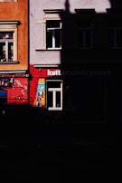 A section of a building facade with vibrant colors is partially obscured by shadows. One part of the building is painted with bright red and contains a single window with a lively mural that includes geometric shapes and colors. Adjacent to it, another section is orange-hued with another window adorned with small objects on the sill.