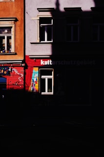 A section of a building facade with vibrant colors is partially obscured by shadows. One part of the building is painted with bright red and contains a single window with a lively mural that includes geometric shapes and colors. Adjacent to it, another section is orange-hued with another window adorned with small objects on the sill.