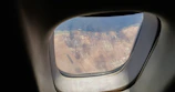 View through an airplane window showing a coastal industrial area. The scene includes various structures, possibly oil tanks and buildings, set against a backdrop of sand and a body of water.