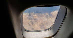 View through an airplane window showing a coastal industrial area. The scene includes various structures, possibly oil tanks and buildings, set against a backdrop of sand and a body of water.