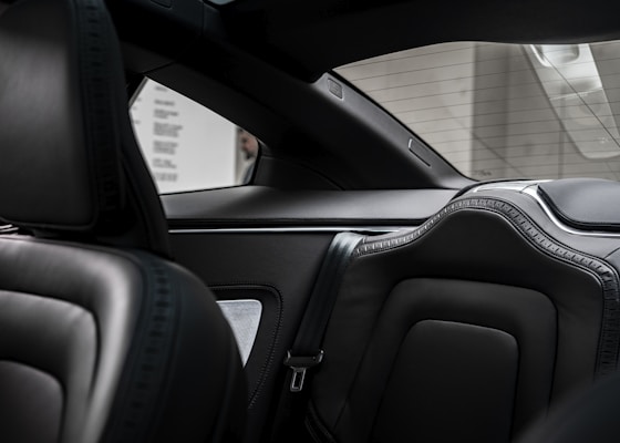 The interior of a luxury vehicle showcasing sleek black leather seats with intricate stitching. The back seat is visible, with detailed padding and stylish design. The view through the back window highlights a parking garage with concrete walls.