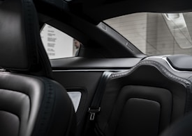 The interior of a luxury vehicle showcasing sleek black leather seats with intricate stitching. The back seat is visible, with detailed padding and stylish design. The view through the back window highlights a parking garage with concrete walls.