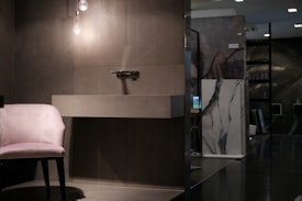 A modern interior scene featuring a sleek, minimalistic washbasin mounted on a textured wall. Below the washbasin, there is soft lighting from a hanging bulb. To the left of the washbasin, a plush pink chair with dark wooden legs is placed. The background includes a marble slab, some artwork, and various pieces of furniture, giving the setting a sophisticated ambiance.