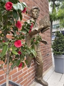 A bronze statue of a man holding a book is integrated into a red brick wall. In front of the statue, a potted plant with vibrant red flowers and green leaves is visible. The background includes a modern urban setting with trees and a partially visible building on the street.