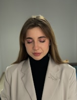A person with closed eyes, wearing a light beige blazer with a Reebok logo and a black turtleneck. The individual has subtle makeup with pink eyeshadow and light lipstick. The background is neutral, enhancing the focus on the person.