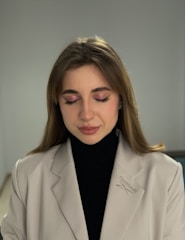 A person with closed eyes, wearing a light beige blazer with a Reebok logo and a black turtleneck. The individual has subtle makeup with pink eyeshadow and light lipstick. The background is neutral, enhancing the focus on the person.
