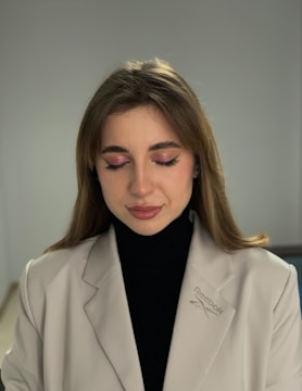 A person with closed eyes, wearing a light beige blazer with a Reebok logo and a black turtleneck. The individual has subtle makeup with pink eyeshadow and light lipstick. The background is neutral, enhancing the focus on the person.