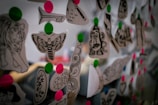 A collection of black and white tattoo designs on paper are pinned with bright pink and green circles on what seems to be a transparent or reflective surface. The designs include images of a moth, dice, a tiger, and a stylized key among others.