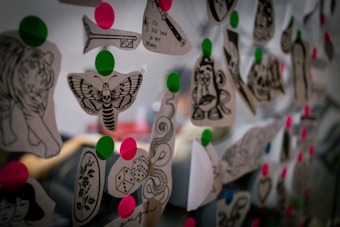 A collection of black and white tattoo designs on paper are pinned with bright pink and green circles on what seems to be a transparent or reflective surface. The designs include images of a moth, dice, a tiger, and a stylized key among others.