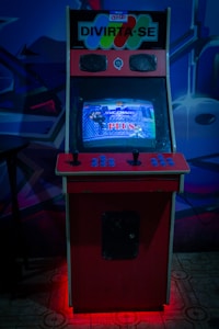 An arcade cabinet with a red and black design is placed indoors. The screen displays a fighting game, and the control panel has joysticks and buttons. The top of the cabinet has a sign with the words 'DIVIRTA-SE' in bold letters.