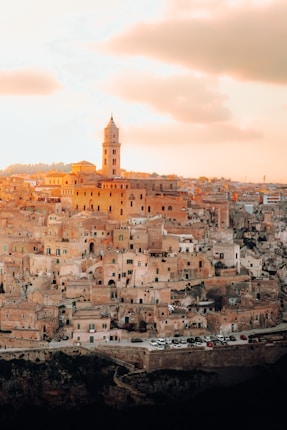 An ancient cityscape bathed in warm, golden light during sunset, showcasing a dense cluster of historical buildings with earthy tones. A prominent bell tower rises above the skyline, contributing to the serene and timeless atmosphere.
